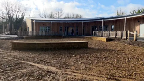 BBC In the foreground is a muddy field with a stone circle in the middle of it. A new wooden clad building curves around the mud.