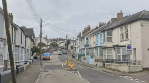 Google image of Clovelly Road in Bideford. Properties line either side of the road. Power cables stretch across both sides of the street. Cars are parked on either side of the road. Vehicles are travelling down the road. 