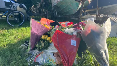 Several bunches of flowers including yellow roses and pink, red and white flowers are laid out on the ground in front of around four boats being stored on a grassed area. 