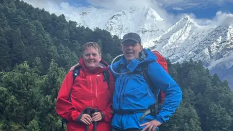 GNAAS This photo shows two hikers standing on a rocky mountain trail surrounded by evergreen forest. They’re wearing outdoor trekking gear - one in a bright red jacket and the other in a blue jacket - with backpacks and hiking poles. Behind them, snow‑covered peaks rise into a partly cloudy sky.