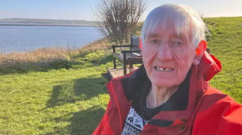 Ray Turnbull is sitting on a bench at Tynemouth, where the river meets the sea. He is wearing a red raincoat and a jumper. The afternoon sun is on the grass beside him and on his grey hair. He is looking straight at the camera and has a sad expression. 