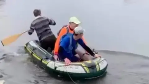 Max Howard The image shows three people in a small inflatable boat on a calm stretch of water. One person at the back is using a single oar to paddle and steer, while the two others sit towards the front of the boat. The boat appears close to the edge of the river or lake, with gentle ripples spreading out around it.