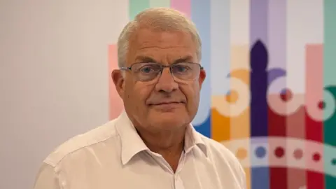 Senator Mark Bolaet is pictured wearing a white shirt and glasses. 
