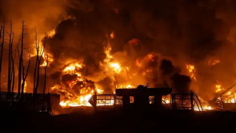 A large fire burns at an oil depot in Iran at night. Flames and thick plumes of dark smoke tower over the outline of a small building.