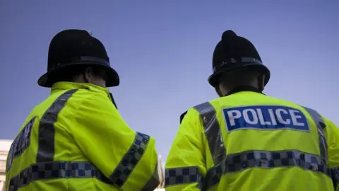 Two police officers are pictured from behind. They are both wearing high visibility jackets with the word 'police' written on the back. They are also both wearing black police hats.