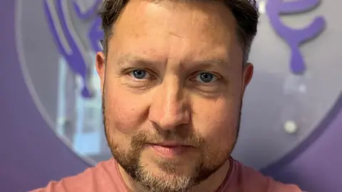 Aaron Ferguson Aaron has short brown hair with white areas and a brown beard with a white patch at his chin. He is wearing a pink/red t-shirt and standing in front of a purple wall.