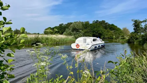 Viewfinder/BBC Weather Watchers A white and blue bloat is travelling across a small stretch of blue water which has long grass and trees either side.
