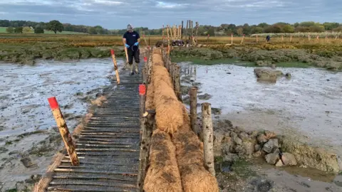 Harwich Haven Authority Another view of the walkway at the saltmarsh were barriers are being made along the right hand side of the walkway. A man wearing welly boots, gloves and a cap stands on it. 