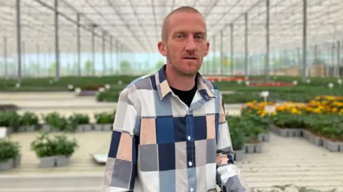 Emily Doughty/BBC Lee Cawood is pictured centre. He has a buzzcut and is wearing a chequered shirt. He is surrounded by punnets of plants, including some which have yellow flowers on them.