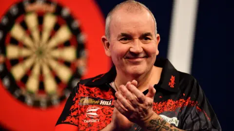 Phil Taylor - a bald man with a red and black sponsored shirt - smiles and claps. A dart board can be seen on a red surface in the background.