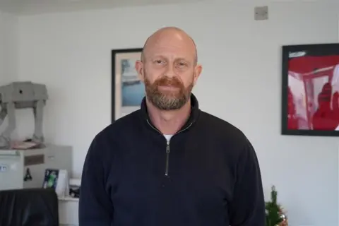 A bald man with a brown beard dressed in a black fleece looks at the camera.