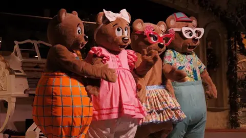 Stephen Adnitt Four people wearing brown teddy bear costumes perform on stage in colourful clothing, with two of them wearing glasses.