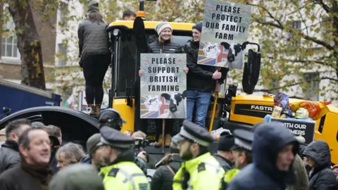 EPA A man and a woman wearing woolly hats stand on a tractor holding signs supporting farmers. They are surrounded by a crowd including members of the police