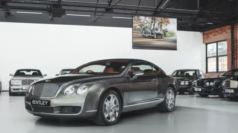 A Bentley showroom with a saloon car in the foreground and other cars lined up against the walls around it. A hanging screen with an image of an older car on a road is hanging from the ceiling behind it.
