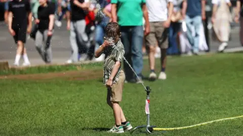 ANDY RAIN/EPA/Shutterstock A boy gets his head wet standing on grass in front of a water sprinkler at Parliament Square in London on 12 July.