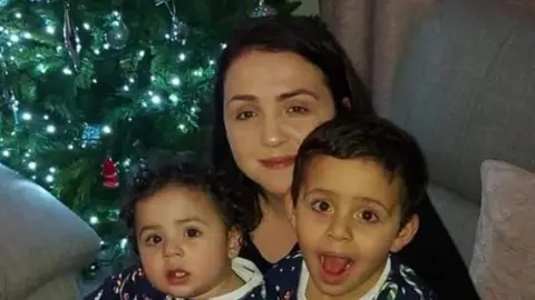 Mandy Kelly A close-up of a woman and two children. They are facing the camera, and there is a Christmas tree behind them. 