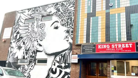 Stu Fern Mural covering the side wall of a two-storey building. It is black and white and of a woman in profile, stylised with patterns, including partial faces, around her. On the right is the entrance to King Street Social Club - marked with a sign above the entrance, and a pattern of blue, white, black and beige, tiles above it.