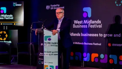 West Midlands mayor Richard Parker is stood at a podium, against a black backdrop with white writing that reads "West Midlands Business Festival". He is wearing a black suit and glasses.