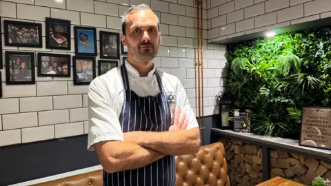 A man dressed in a white shirt and a blue chef's apron stands with his arms folded next to a table and brown chair in a restaurant. Behind him are framed pictures mounted on a white-tiled wall. To his left is a large plant.