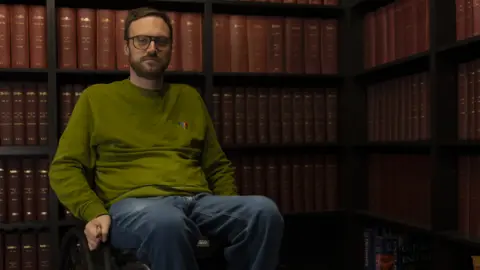 Matthew Bassett who has short brown hair, glasses and a beard and is wearing a long-sleeved green t shirt and blue jeans. He is sat in his wheelchair, in front of a bookcase full of brown books.