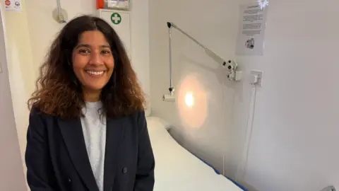 BBC/Jim Scott Dr Nousa Ali, who is wearing a blue blazer on top of a grey shirt, stands in front of a patient bed inside her GP practice. In the background is a light, and a first aid box.