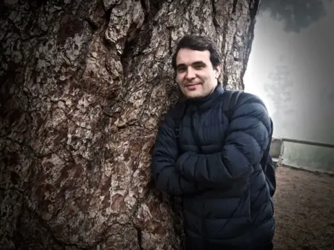 A man in a black jacket stands next to a tree, smiling