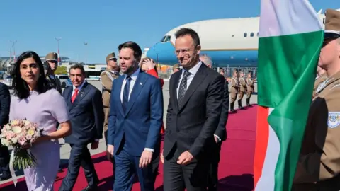 Reuters A woman in lilac with flowers walks alongside two men in suits beside a flag