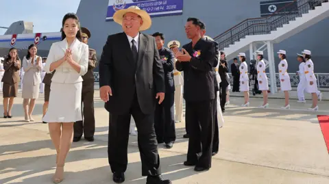 Kim Jong Un and his daughter Kim Ju Ae walk past honor guards with a destroyer in the background