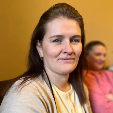 Leeann is sitting and she is looking into the camera. One of her daughter's is sitting behind her.