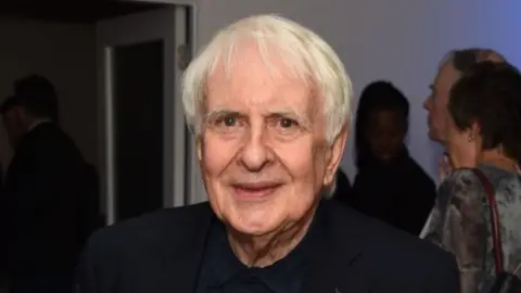 A man with white hair looks at the camera