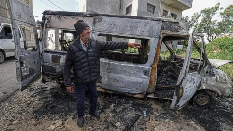 AFP via Getty Images An elderly man stands next to a burnt-out van. 