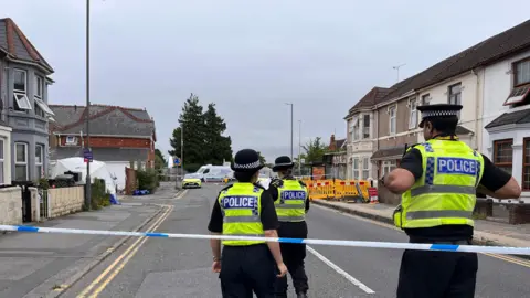 Three police officers stood by police tape inside a police cordon. They are wearing high vis jackets and black police hats. You can see houses in the background.