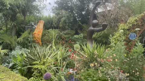 Katie Jaffar An exotic front garden can be seen. It is filled with sculptures, plants, flowers and trees.