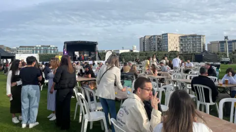 People are sitting at wooden tables in a field with a stage in the background with flats behind that.