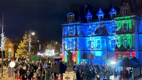 Hanley Town Hall, illuminated with blue and green lighting, with a crowd of people gathered outside it with a stage and gazebo to one side.