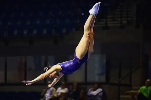 British Gymnastics Akbar competing in gymnastics in mid-air arms outstretched in purple leotard