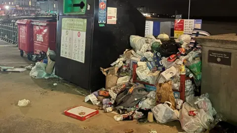 Kieran Hunt Mixed rubbish including cardboard, food packaging and bags overflowing onto the ground beside a recycling station in Margate