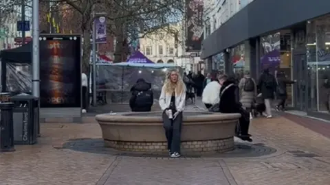 Georgia Pasco A woman sitting in the middle of a city centre while singing.