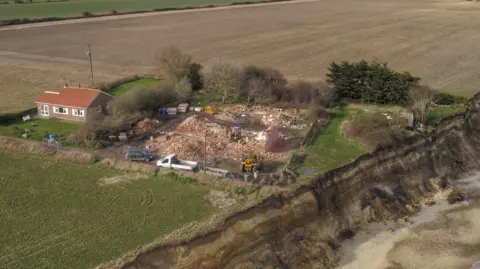 Martin Barber/BBC A pile of rubble following the demolition of a house. The site is by a sandy cliff edge and is backed by a field. A bungalow sits next to it. 
