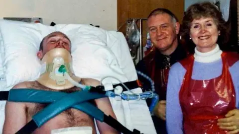 Family photo A topless man with a neck brace lying in a hospital bed. His hairy chest is strapped to the bed by blue cross-ver straps. Beside him are a man and woman. They both have dark hair and the man has an arm around the woman. He is wearing a dark shirt and sge is wearing a lilac jumper with a red plastic apron over it. They are both smiling and looking at the camera.