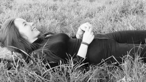 FEES Fund A black and white photo of Fiona Braidwood laying down in tall grass. She has long straight hair and is wearing black skinny jeans and a long-sleeved black top with a high neck. She is laughing with her eyes closed and her hands clasped together in front of her waist.