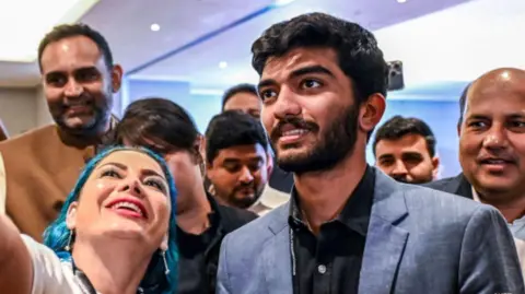 Getty Images Gukesh poses for a selfie with chess fans, wearing a blue blazer and black shirt. He is standing next to a woman and is surrounded by people behind.