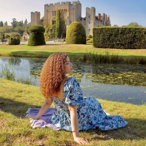 @eboniivoryblog Eboni, a woman in her 30s with curly brown hair, sitting on the grass of a riverbank wearing a blue and white floral puff sleeved dress and looking across the river at Hever Castle in Kent