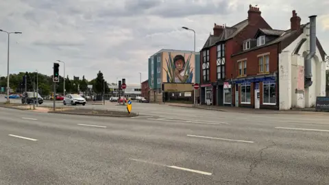 A busy road in Carlisle with multiple lanes and a traffic light to the left. There are cars coming down the right side towards the camera. Behind the road are two large red brick buildings, with another building to the left of them with a mural of a young boy on the side. There are trees in the distance and the sky is covered in white and grey clouds.