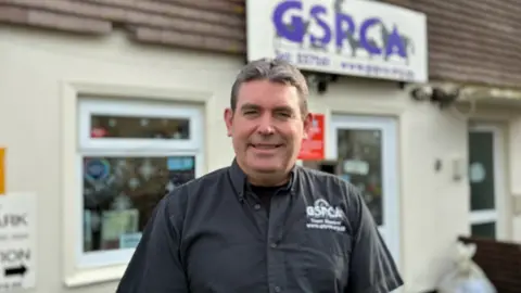 Steve Byrne facing the camera, infront of a large GSPCA sign. He has short dark hair, and is wearing a button up shirt with the GSPCA logo on it.