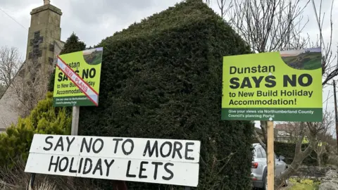 A wall covered in ivy has three signs in front saying Say No to More Holiday Lets. Two are green, professionally designed and on wooden poles. One is painted black letters on a white background.