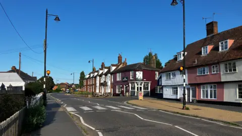 Station Road in Woodbridge. A pedestrian zebra crossing can be seen with a pub in the distance and row of houses.