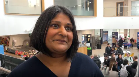 Rakhee Aggarwal, a woman with a black bob who is wearing a dark v-neck top, stands above university students at a careers fair