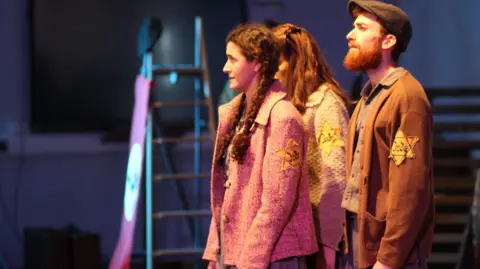 Voices of the Holocaust Three characters stand on stage acting a scene from Kindness. Susan's character is at the front. She has long plaits and wears a pink jacket and a yellow star on the arm, saying "Jude" - German for "Jew.". The others wear the same star. A man with a beard and cap is to Susan's left and another woman to her right. There is a step ladder with a Nazi flag in the background.