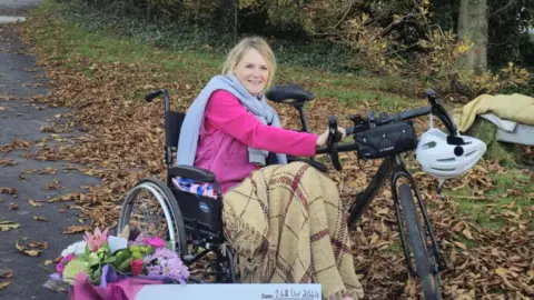 Family handout Alison Warner sits in a wheelchair and is smiling with her hands on a bike. 